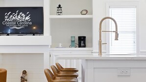 Une cuisine moderne avec un plan de travail blanc, un robinet en acier inoxydable, un tabouret de bar et un téléviseur diffusant des publicités pour des « séjours et des offres spéciales en Caroline du Nord, sur la côte ».