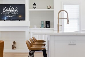 A modern kitchen with a white countertop, a stainless steel faucet, a bar stool, and a television displaying 'Coastal Carolina Vacations and Sales'.