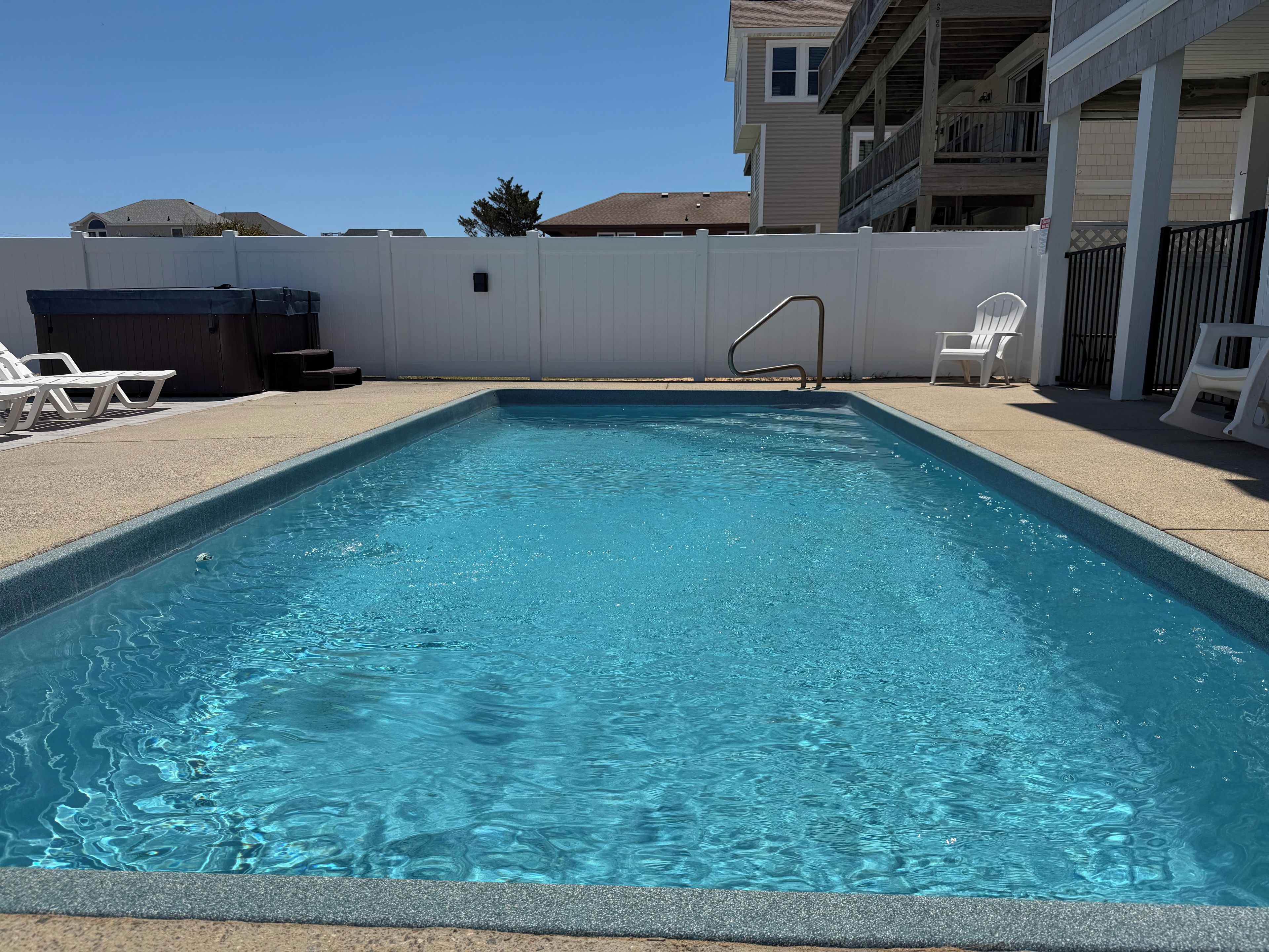 Une piscine rectangulaire avec un escalier en métal, entourée d’une clôture blanche, et des chaises longues blanches disposées d’un côté.
