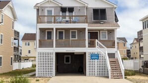 Une maison de deux étages située en bord de mer, dotée d’un balcon, d’un porche et d’une enseigne indiquant « Coastal Carolina Vacation Rentals and Sales » (locations et ventes de résidences de vacances à Coastal Carolina).