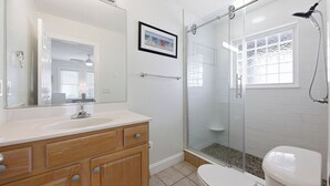 Un baño con ducha envuelta en vidrio, inodoro blanco y tocador de madera con espejo.