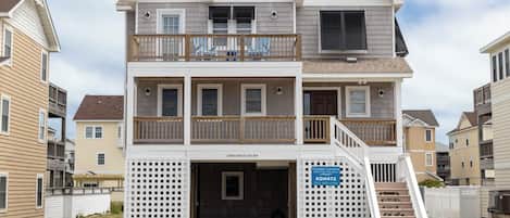A two-story beachfront house with a balcony, a covered entryway, and a sign indicating Coastal Carolina Vacation Rentals and Sales.