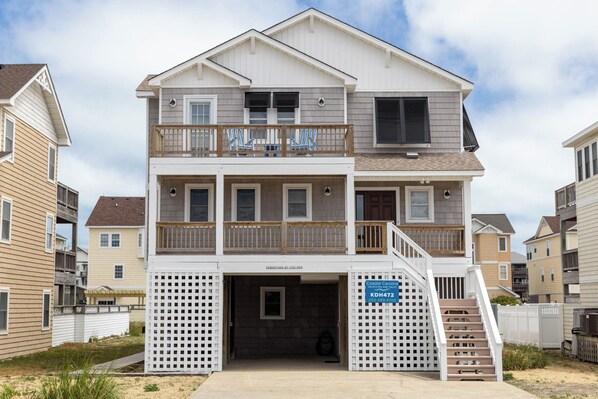 A two-story beachfront house with a balcony, a covered entryway, and a sign indicating Coastal Carolina Vacation Rentals and Sales.