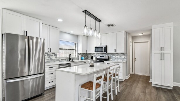 A modern kitchen with white cabinetry, stainless steel appliances, and a central island with bar stools. - Coastal Family Retreat – Heated Pool, Fire Pit & Fun! (Holiday)