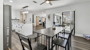 A modern kitchen with white cabinets, a central island, and a dining area with a table and chairs. - Coastal Family Retreat – Heated Pool, Fire Pit & Fun! (Holiday)