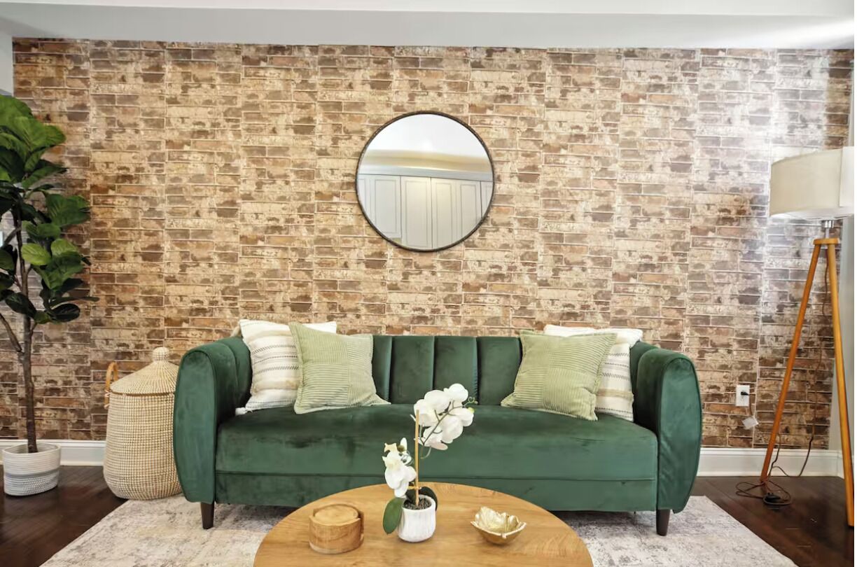 A green sofa with white and green cushions, a round mirror on the wall, a potted orchid, and a wooden coffee table.