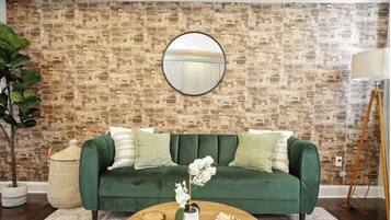 A green sofa with white and green cushions, a round mirror on the wall, a potted orchid, and a wooden coffee table.