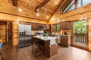 A wooden cabin kitchen with a central island, stainless steel appliances, and a large window overlooking greenery.