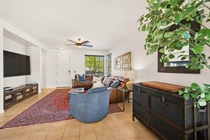 A living room with a sofa, a coffee table, a television, a ceiling fan, and a large plant. - Heated Pool Oasis | Family Retreat in Gilbert (Gilbert)