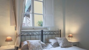 A bedroom with a bed, two bedside lamps, a window with curtains, and a view of buildings outside.