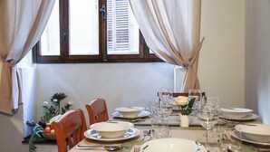 A dining table set for four with white plates, cutlery, and glasses, a vase with flowers, and a window with curtains.