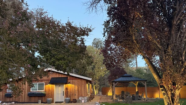 Una cabaña rústica con camino de entrada de grava, fachada de madera y un área de estar al aire libre techada.