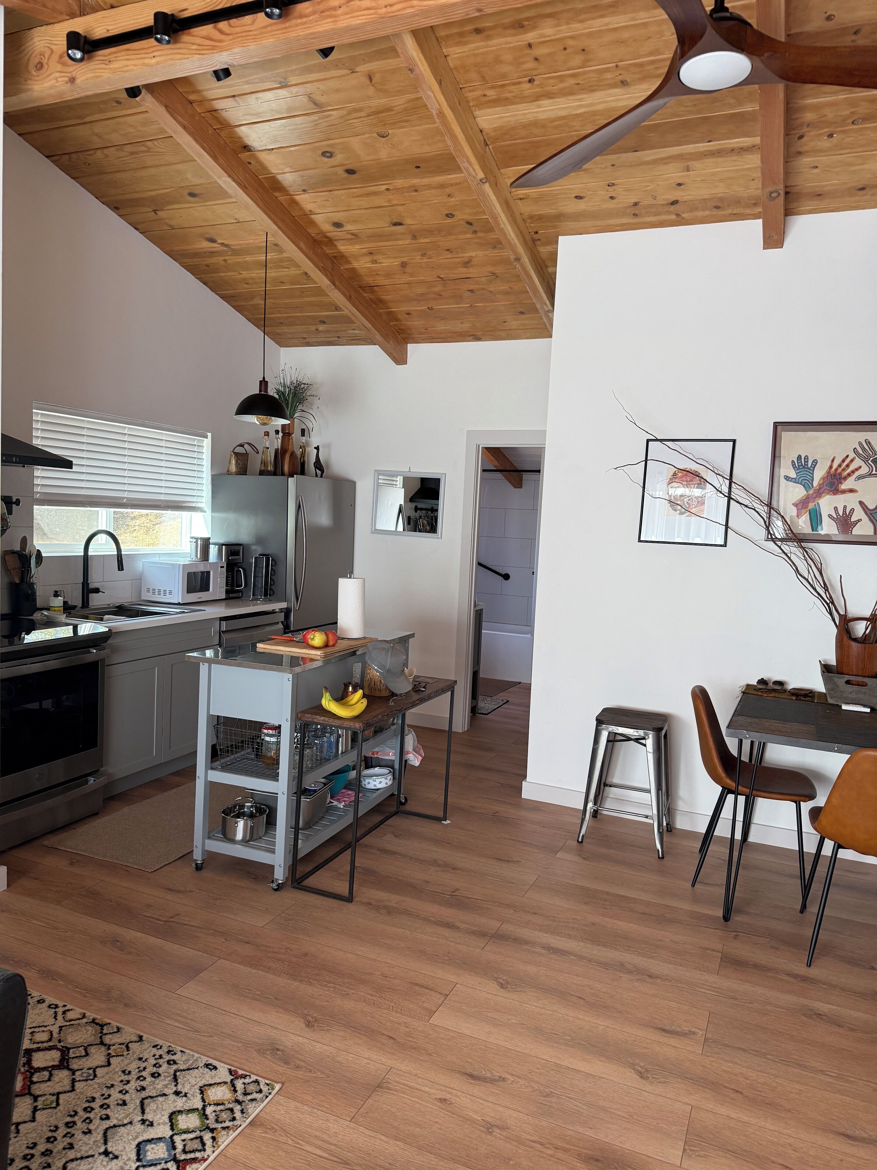Une cuisine moderne avec des poutres en bois au plafond, un coin repas avec une table et des chaises, et un petit chariot de cuisine contenant des fruits et des bocaux.