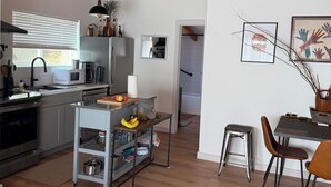 Una cocina moderna con vigas de madera en el techo, un comedor con mesa y sillas, y un carrito pequeño con frutas y frascos.
