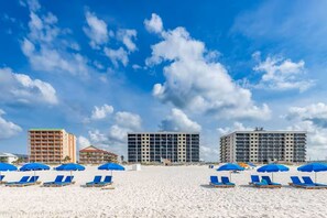A beach with blue umbrellas and lounge chairs, a hotel building, and a clear blue sky.