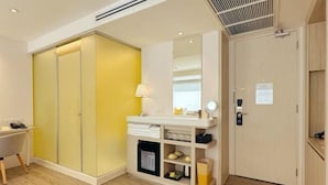 Een hotelkamer met een houten vloer, een wit bureau met stoel, een geel kledingkastje, een kaptafel met spiegel, en een deur met slot.
