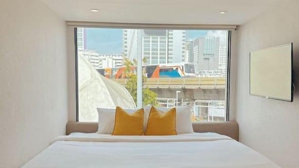 Een hotelkamer met een groot raam, een bed met witte beddengoed en twee gele kussens, en een aan de muur bevestigde televisie.