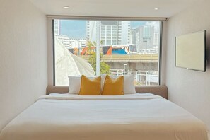 A hotel room with a large window, a bed with white bedding and two yellow pillows, and a mounted TV on the wall.