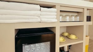 Een netjes opgeruimde hotelkamer met een plank waarop opgevouwen handdoeken, glazen potten en een zwart apparaat, mogelijk een koffiezetapparaat, staan.