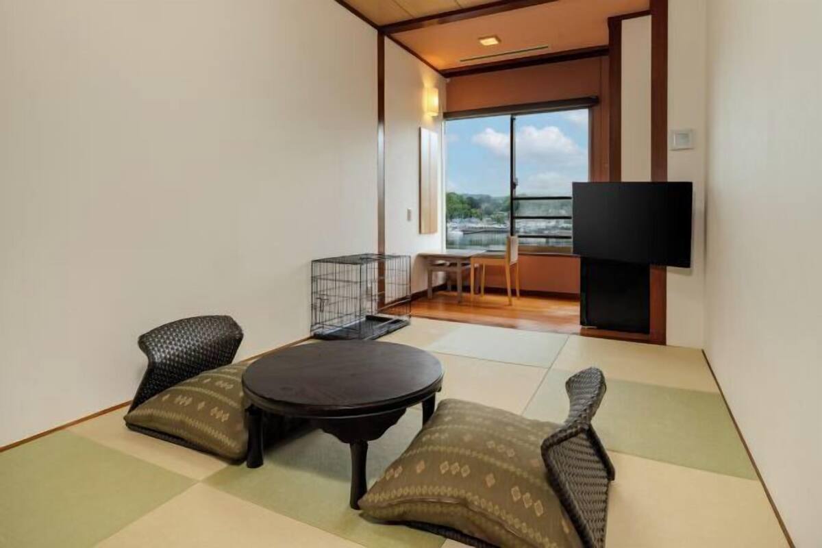 Una habitación con suelo de tatami, una mesa baja, dos sillas con cojines, un televisor y vistas a un edificio por la ventana.