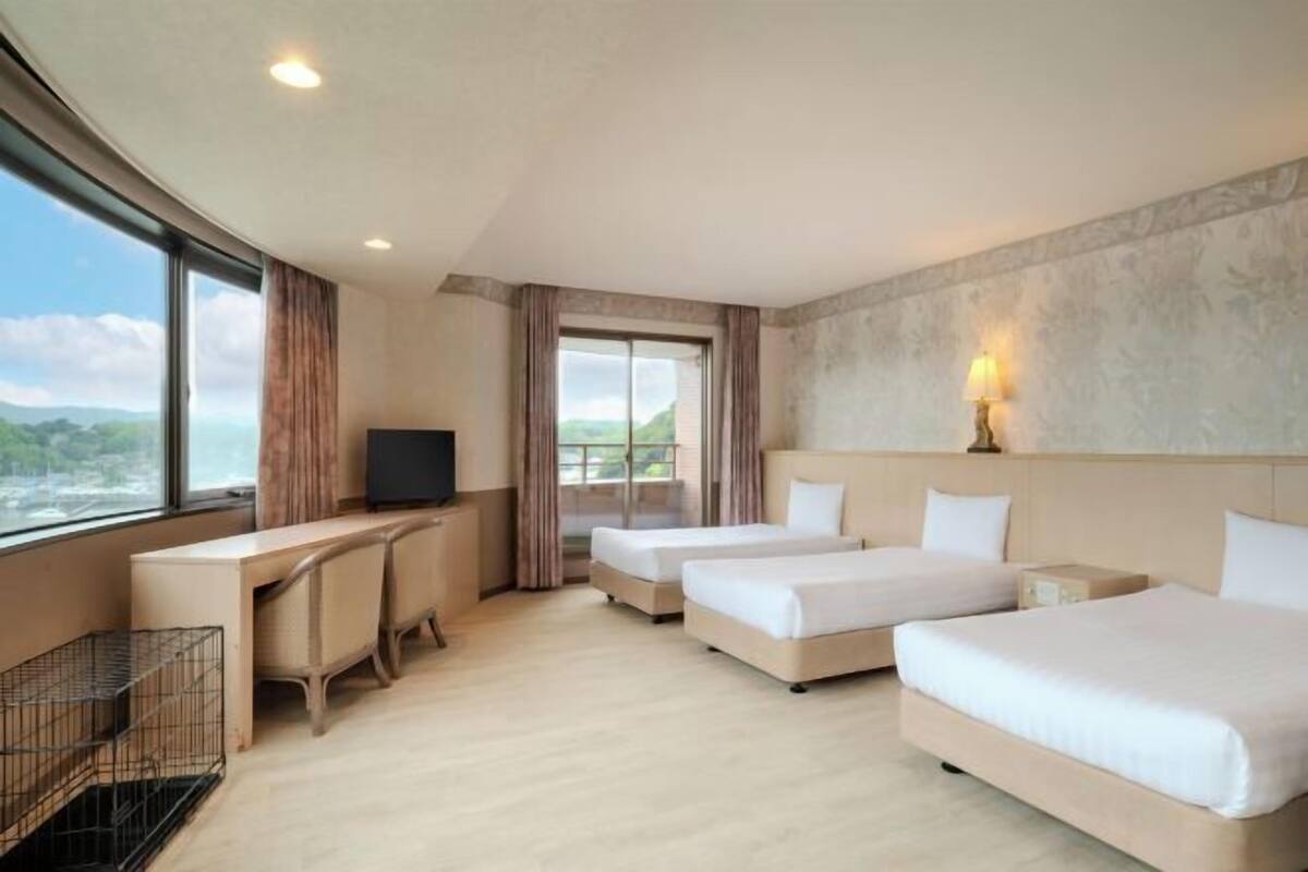Habitación de hotel con escritorio, dos camas, televisor, lámpara y vistas al exterior.