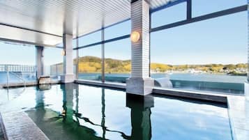 Piscina interna com paredes de vidro transparente, vista para um corpo d'água e paisagem ao longe.