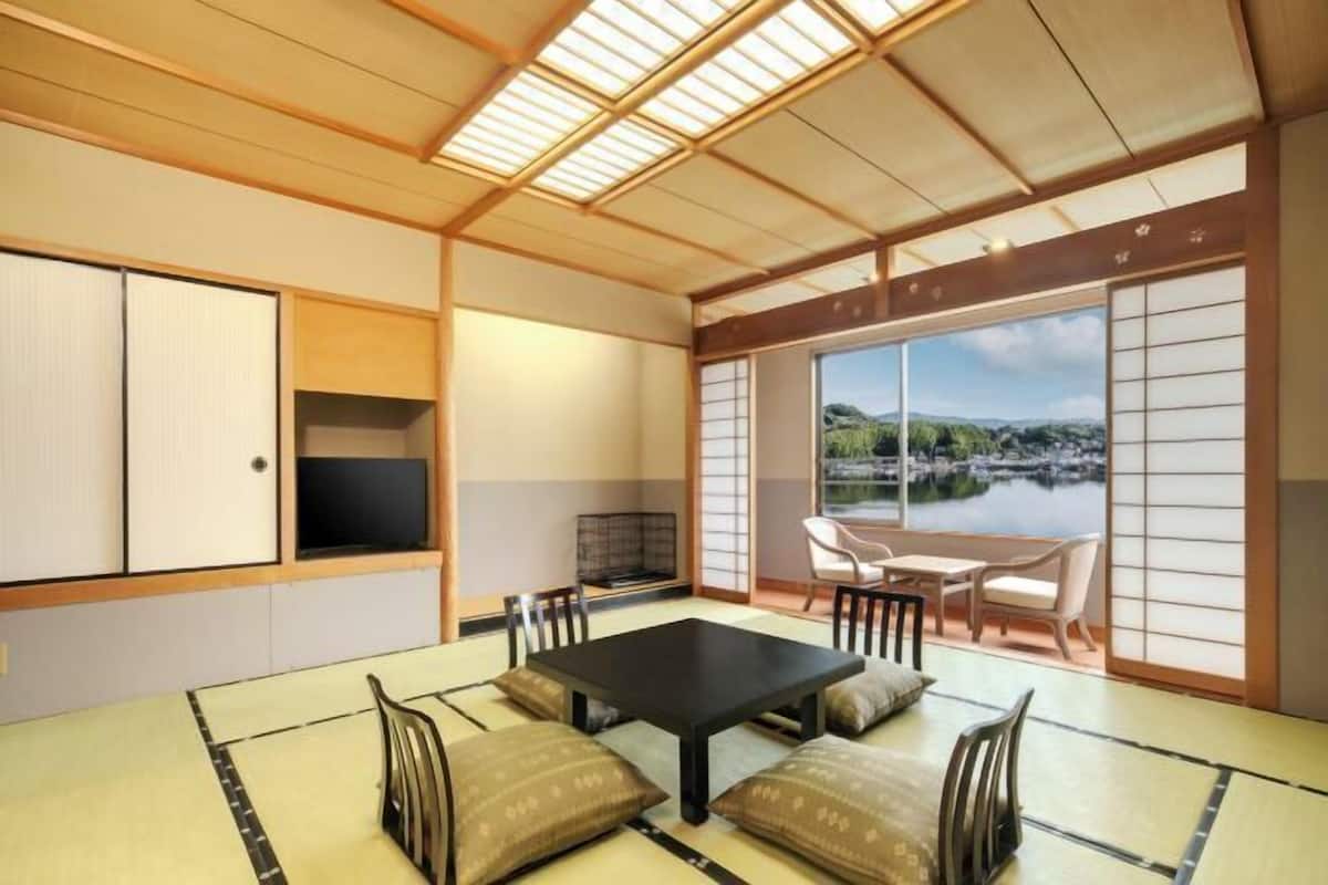Una habitación japonesa tradicional con suelo de tatami, una mesa negra, sillas, un televisor y vistas a un lago.