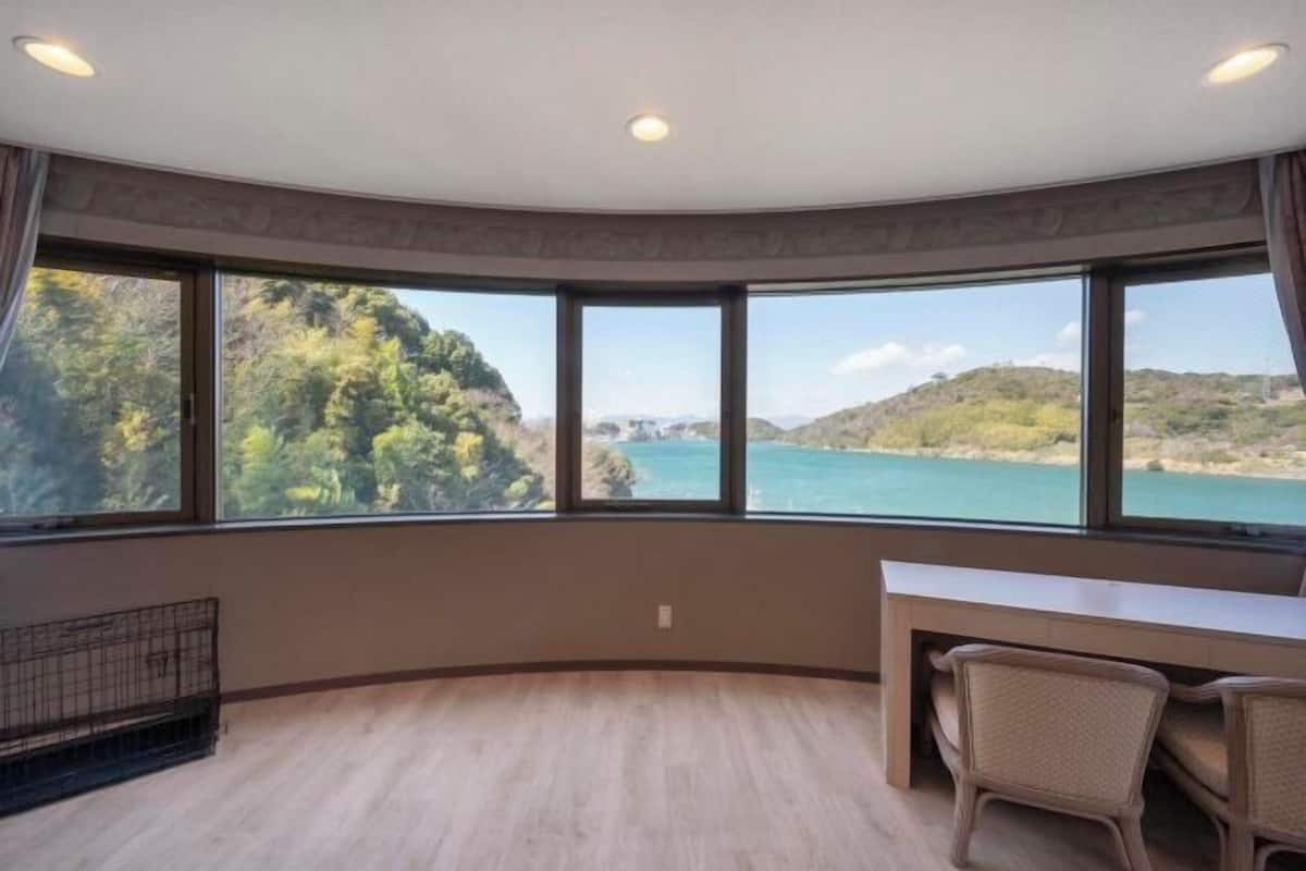 Una habitación con techo curvo, ventanas amplias con vistas a un paisaje costero, un escritorio con sillas y una pequeña zona cerrada con una reja de metal.
