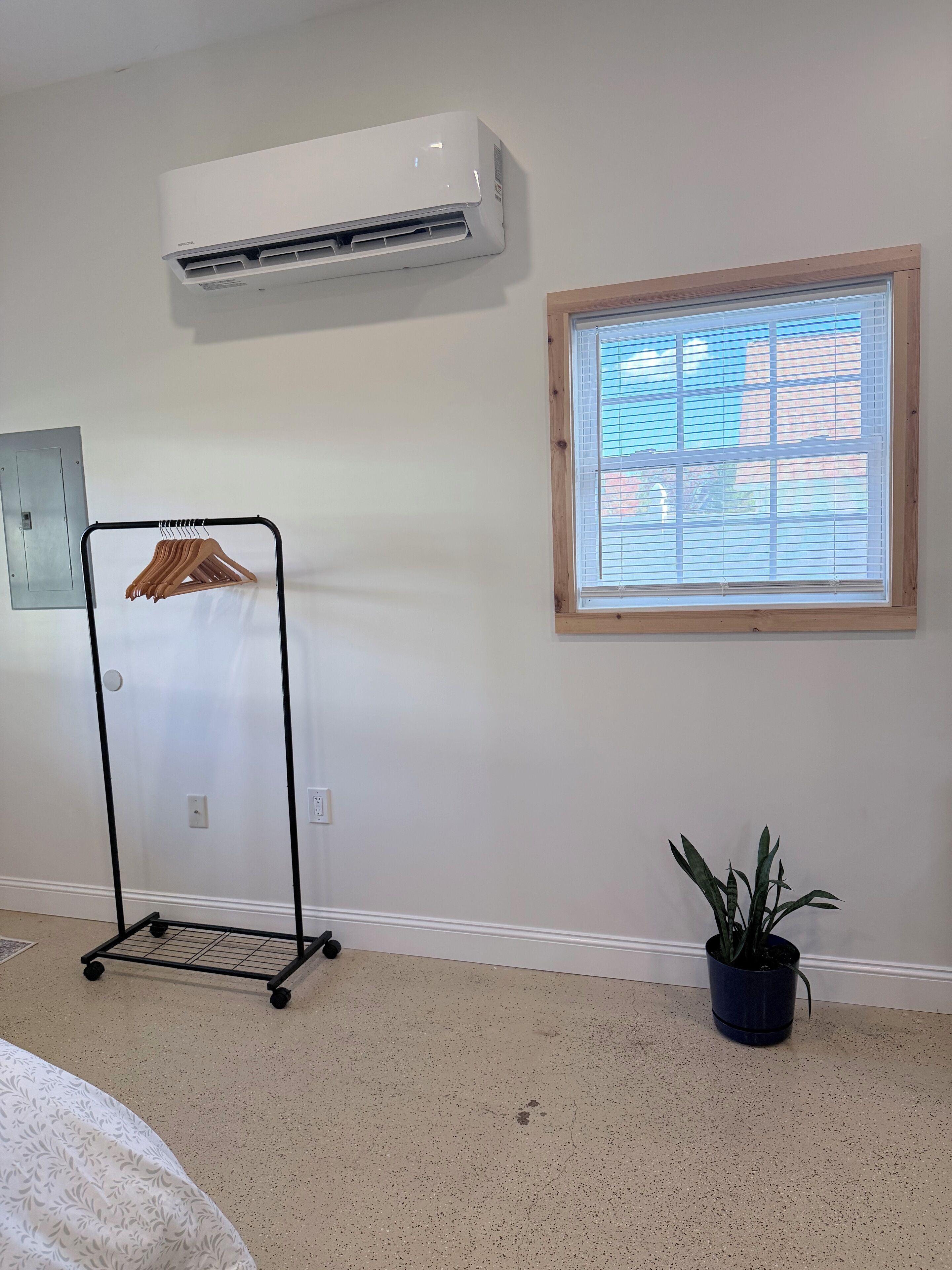Une pièce avec un climatiseur blanc, une fenêtre avec des stores, un portant métallique avec des cintres et une plante en pot.