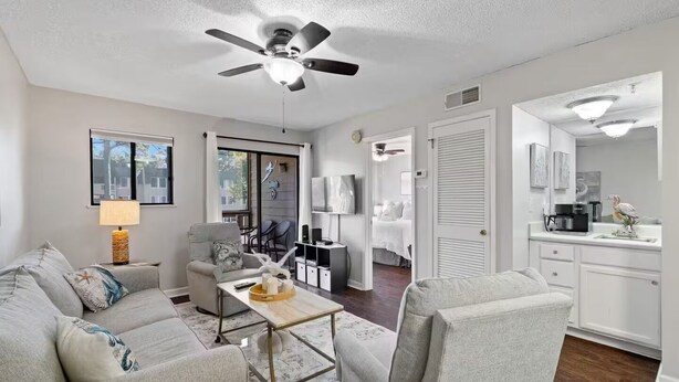 A modern living room with a grey sofa, a coffee table, and a ceiling fan. There is a kitchen area with white cabinets and a countertop.
