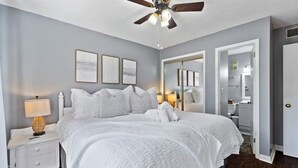 A neatly made bed with white linens, a bedside lamp, and a ceiling fan in a room with grey walls and a wooden floor.