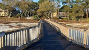 A wooden boardwalk leading to a building with multiple balconies, surrounded by trees and open grassy areas.