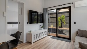 A modern hotel room with a bed, a TV, a small table, a sofa, and a sliding glass door leading to a balcony with a wooden fence and greenery.