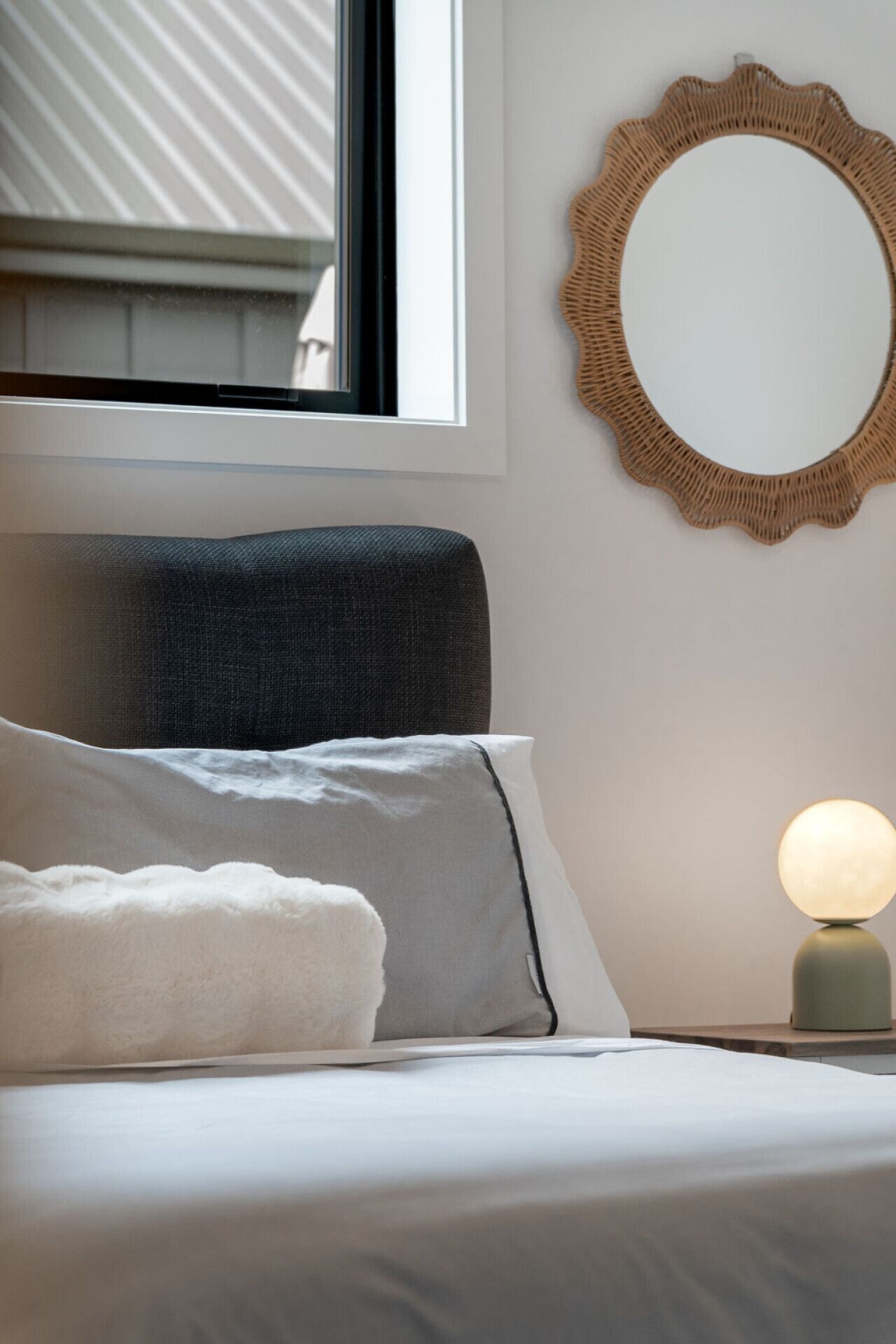 Una cama bien hecha con ropa blanca, cabecera gris y almohada blanca. Hay un espejo redondo tejido en la pared y una mesita de noche con una lámpara junto a la cama.