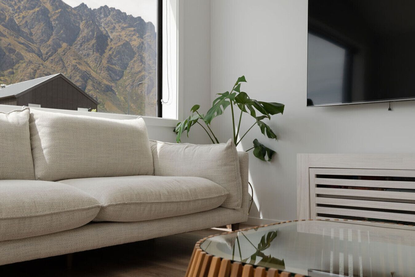 Uma sala de estar moderna com um sofá bege, uma planta grande e vista para montanhas pela janela.