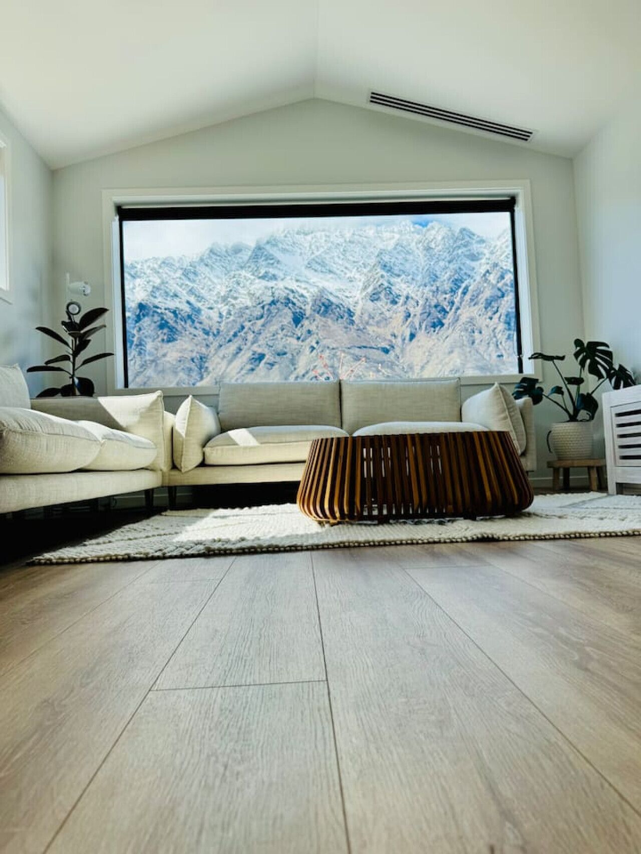 A modern living room with a large window, a beige sofa, a wooden ottoman, and a view of snow-capped mountains.