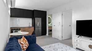 Una sala moderna con un sofá azul oscuro, un mueble blanco para televisión y una zona de cocina con armarios oscuros y un refrigerador.