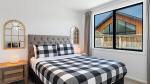 Un dormitorio con una cama grande, dos mesitas de noche, una ventana con vistas a una casa de madera y dos espejos adosados a la pared.