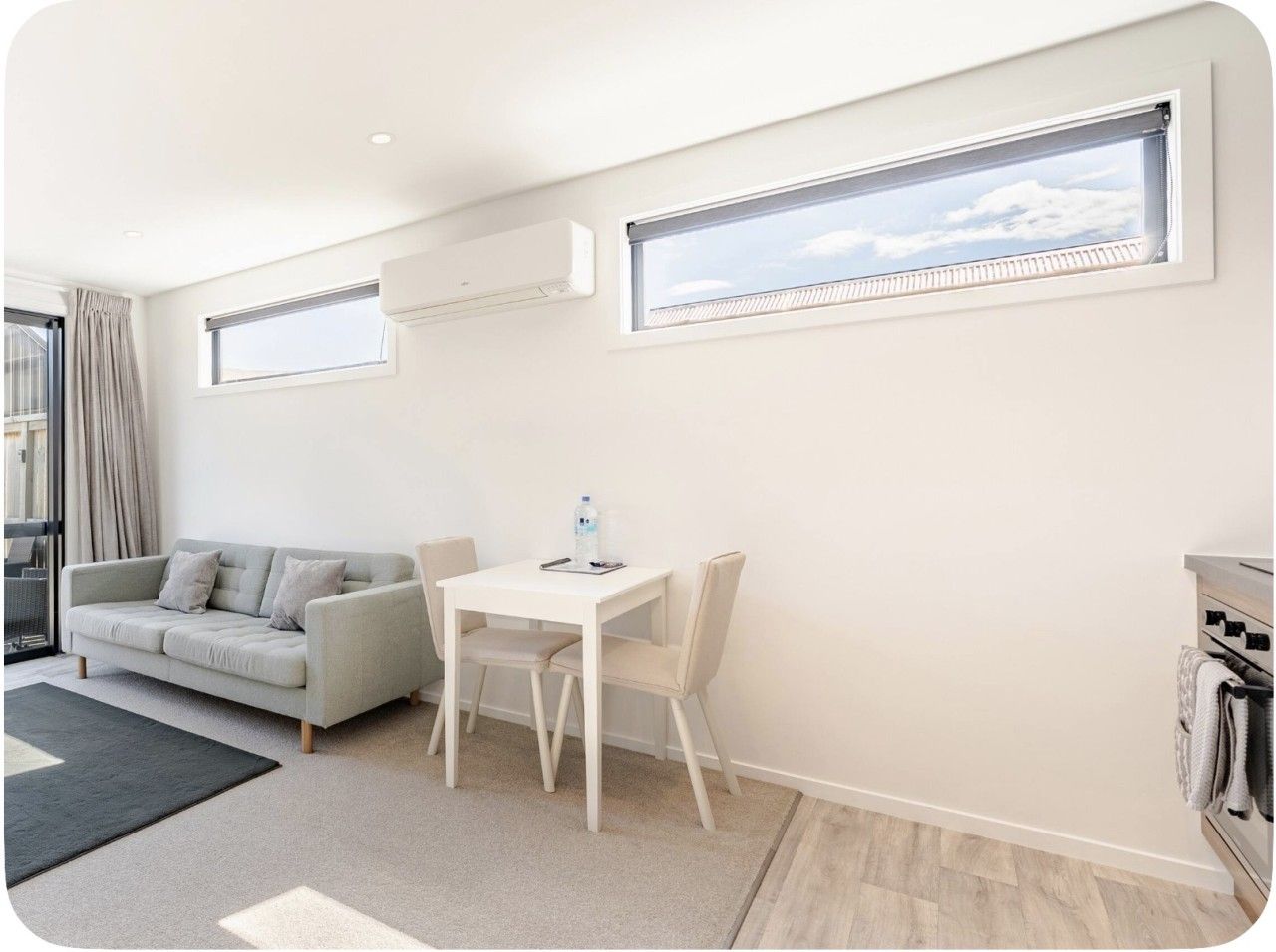 A modern living room with a sofa, a dining table, and chairs. There are two windows with blinds, a ceiling-mounted air conditioner, and a view of the sky.