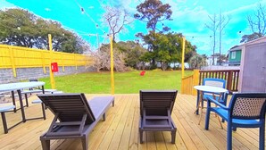Eine Holzterrasse mit Gartenmöbeln, darunter Liegestühle und ein Tisch, mit Blick auf einen Rasen mit Bäumen und einem Gebäude im Hintergrund.
