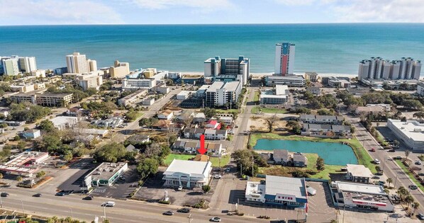 Une vue aérienne d’une ville côtière, avec un mélange de bâtiments résidentiels et commerciaux, un hôtel imposant sur lequel pointe une flèche rouge, et une vaste étendue d’eau en arrière-plan.