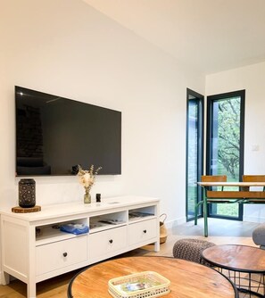 Una sala de estar moderna con un armario blanco, un televisor de pantalla plana, una mesa redonda de madera y un banco junto a un ventanal con vistas a zonas verdes.