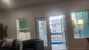 Een moderne woonkamer met een zwarte leren bank, een glazen salontafel en een deur naar een balkon met uitzicht op een woonwijk.