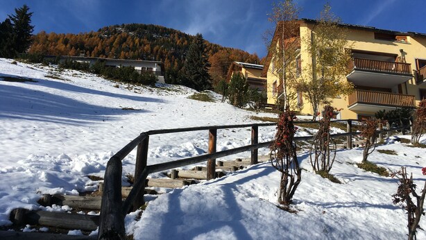 Un paysage enneigé avec une clôture en bois, un bâtiment jaune doté de balcons et une montagne en arrière-plan.