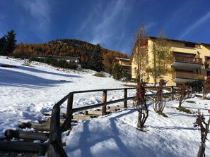 A snow-covered landscape with a wooden fence, a yellow building with balconies, and a mountain in the background. - Amazing 1-bedroom apartment in Samedan (Samedan)