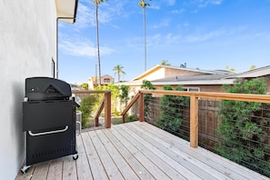A wooden deck with a barbecue grill, a railing, and a view of a residential area with houses and palm trees.