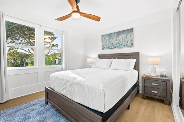 A bedroom with a bed, a nightstand, a lamp, a ceiling fan, and a window with a view of trees.