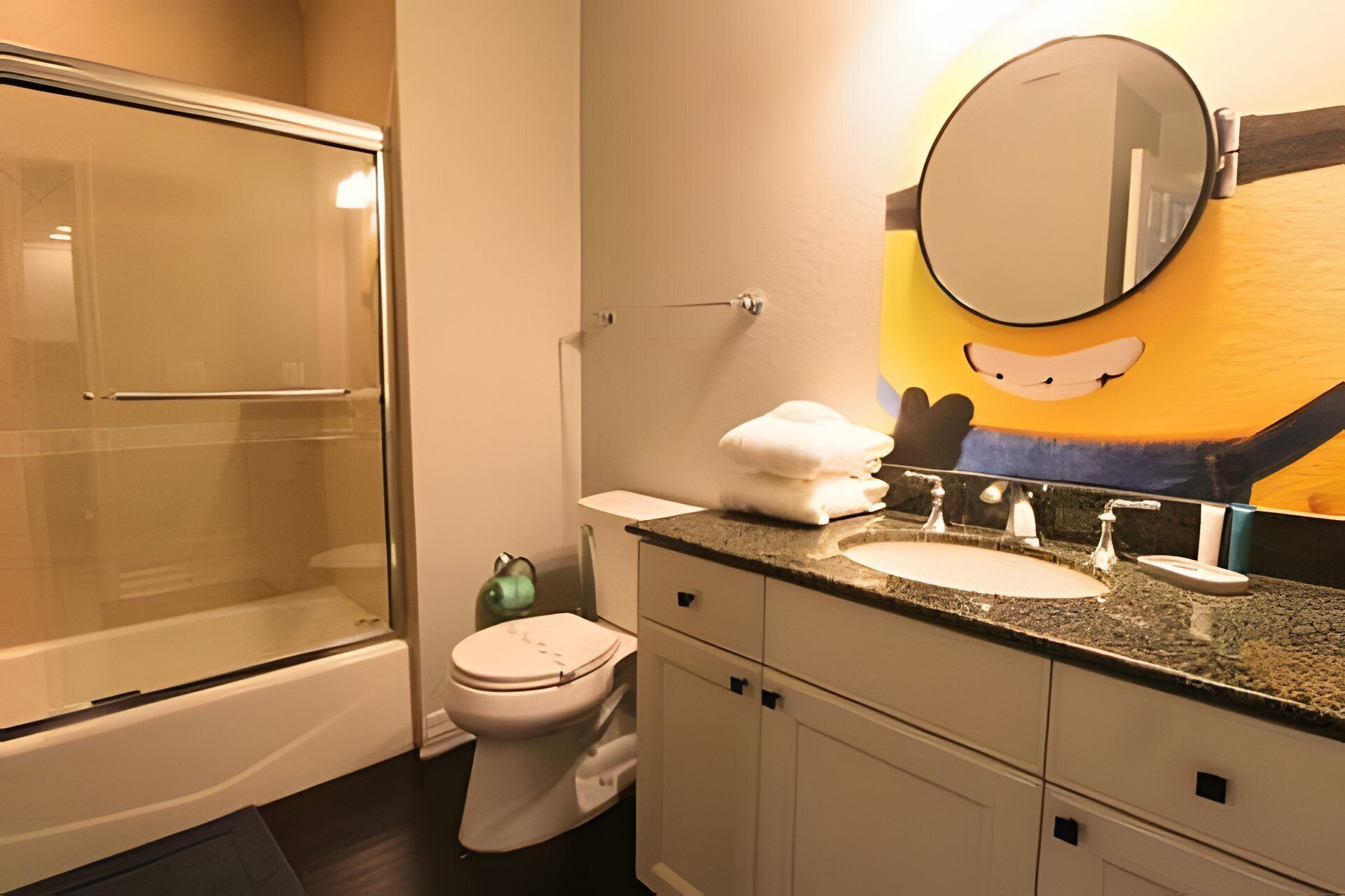 A bathroom with a glass shower enclosure, a toilet, a sink with a mirror, and a cartoon character on the wall.