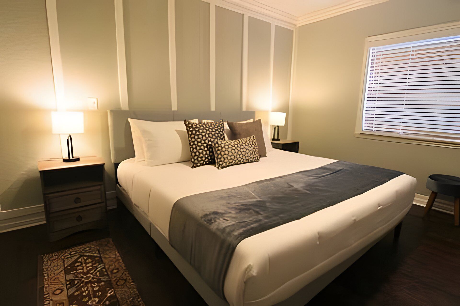 A neatly arranged bedroom with a large bed, two bedside tables, a nightstand, a window with blinds, and a patterned rug.