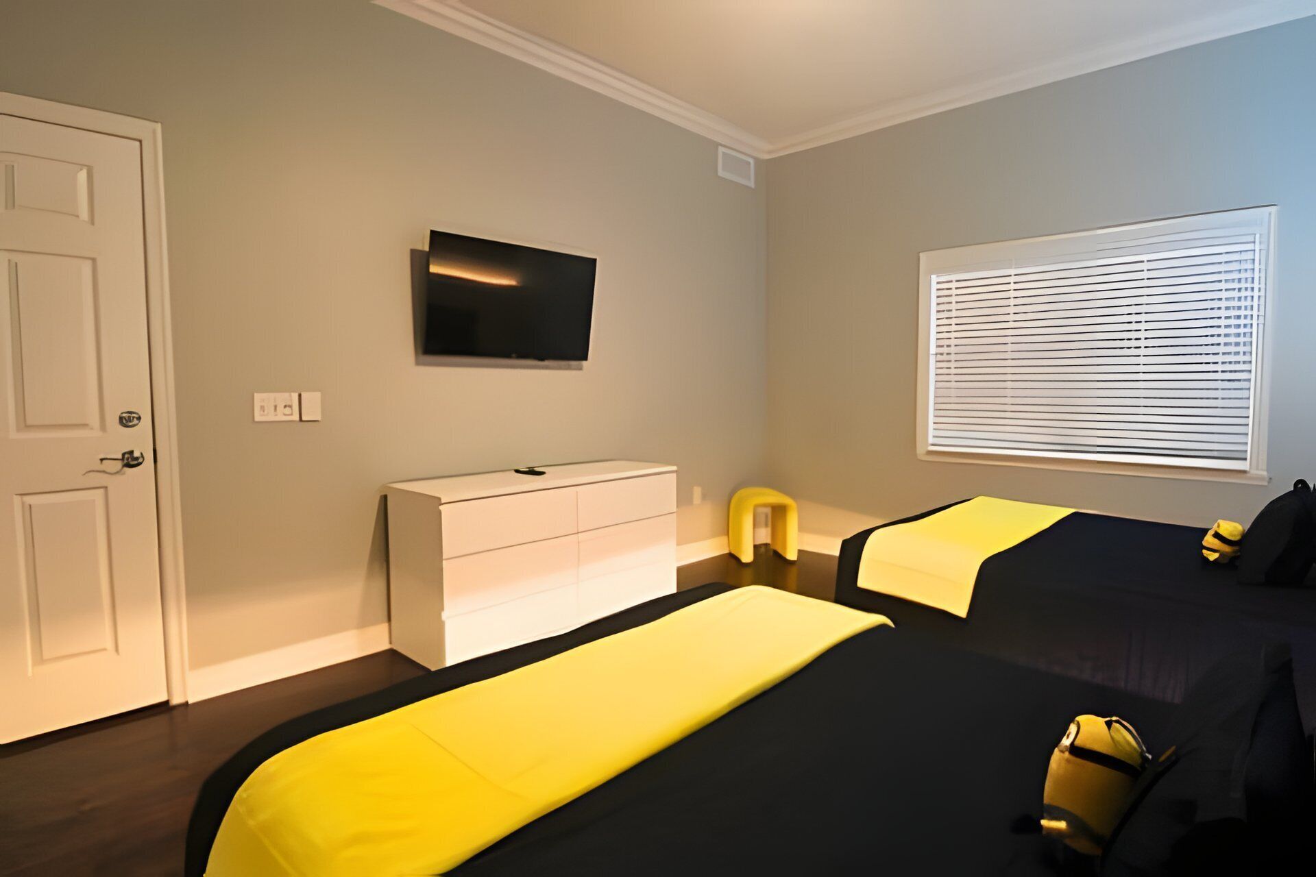 A hotel room with two beds, a TV mounted on the wall, a window with blinds, and a white dresser.